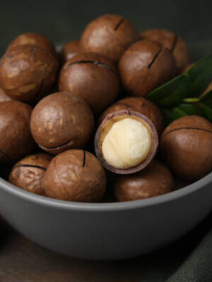 Tasty macadamia nuts in bowl and green twigs on table, closeup